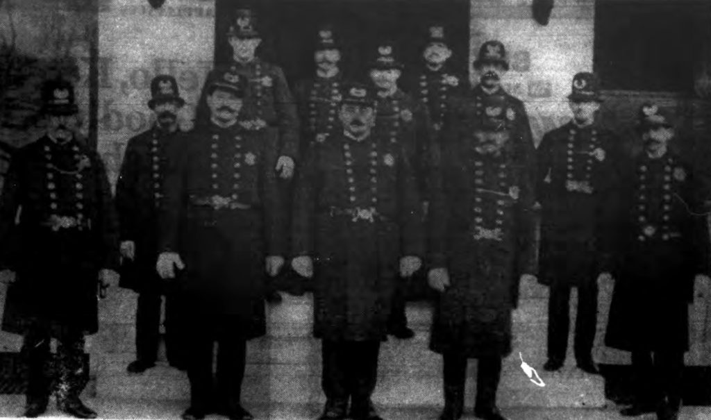 "FOURTH PRECINCT POLICE—Above is the Lansingburgh police force as it appeared in the early 1900s, a few years after Lansingburgh became a part of Troy.  The picture was taken in front of the precinct station house on the east side of Second Avenue between 116th and 117th streets.  Can you name those not identified?  Sgt. Albert D. Chesbrough is in center of front row, Sgt. William M. Flynn, second left, and Ptl. Holman P. Shires at extreme left.  Between Flynn and Shires on the step is Ptl. Samuel E. Rounds.  Behind Flynn is Ptl. James O'Brien who later became captain of the precinct.  The officer standing behind Sergeant Chesbrough's left shoulder is Ptl. Louis Hansen.  The man, front row, second right is believed to be Ptl. Daniel Bird, and between him and the officer on extreme right is either Ptl. Joseph Schultz, who also later captained the precinct, or Ptl. John L. Rabie. (Photo loaned by George MacCulloch)" The Record. October 5, 1968: B22.