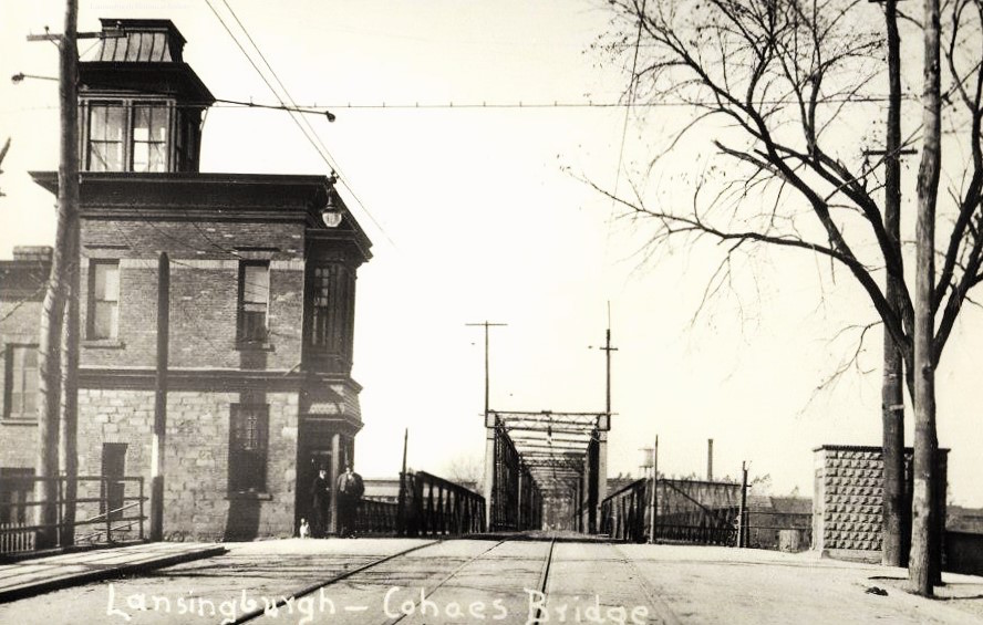 View facing west at approach to bridge, showing rail tracks, two-story brick gatehouse topped by a third-story cupola on south side with two men and a dog standing at its door, and a small brick structure of some kind on the north side.  A water tower can be seen in the distance on the north side of Van Schaick Island.