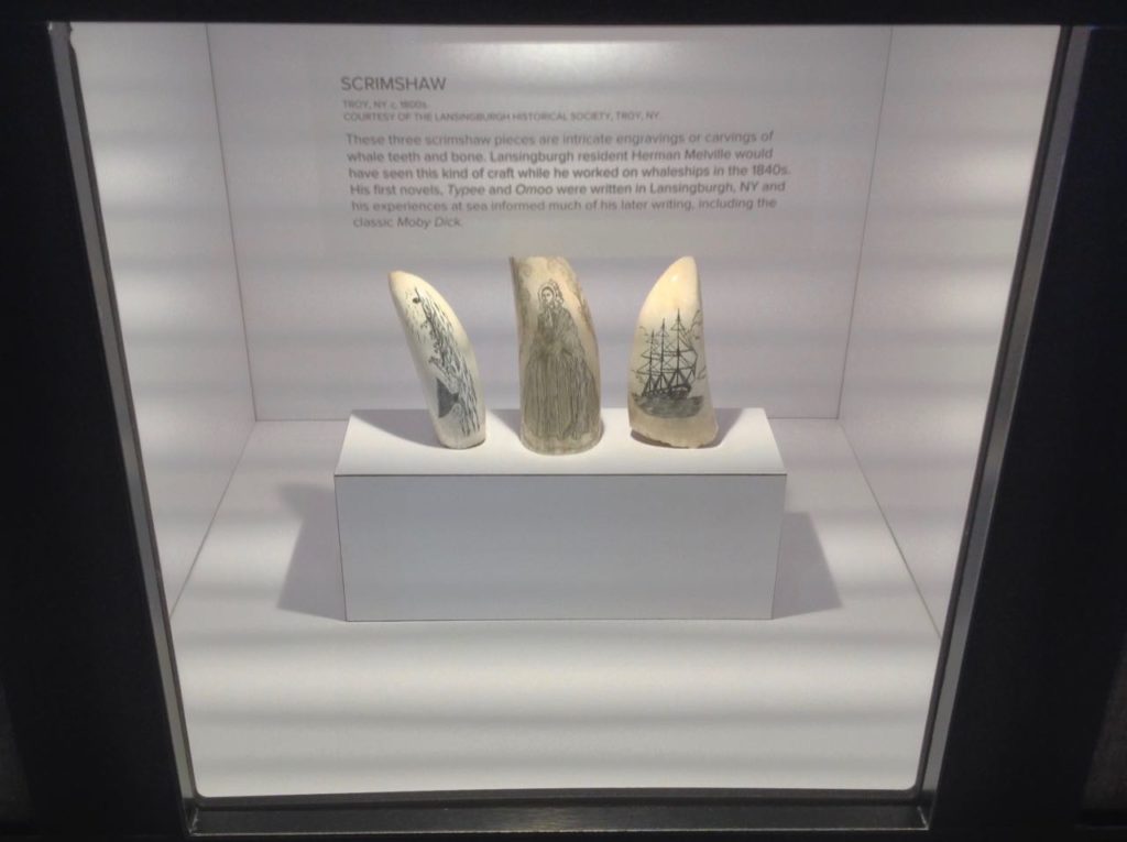 Three pieces of modern engraving on whale's teeth in a lighted cabinet, one of a whaling scene, another of a woman in fancy clothing, another of a whaling ship.